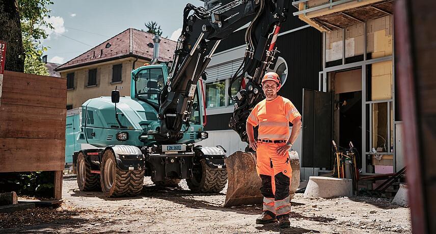 Pracovník v reflexním pracovním oděvu stojící vedle kolového rypadla Wacker Neuson EW100 na městské stavbě.