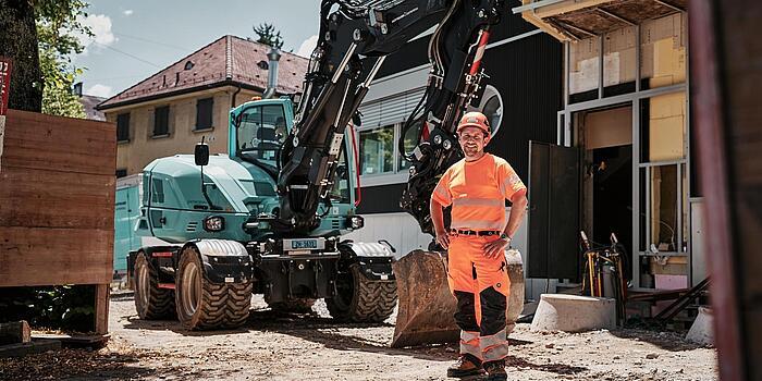 Operator wearing high-visibility workwear standing beside a Wacker Neuson EW100 wheeled excavator at an urban construction site.