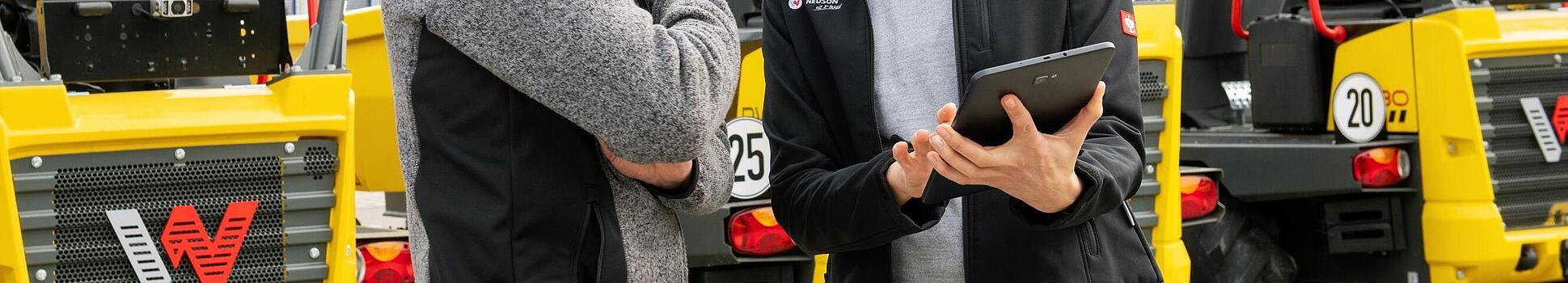 Usměvavý zaměstnanec Wacker Neuson s tabletem radí usmívajícímu se zákazníkovi před stavebními stroji Wacker Neuson.