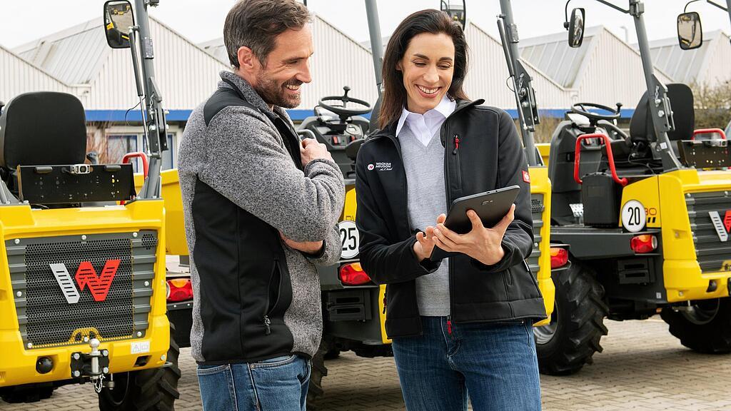 Smiling Wacker Neuson employee with tablet advising smiling customer in front of Wacker Neuson construction machines.