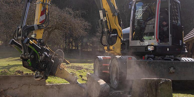 Kolové rypadlo Wacker Neuson EW100, které na lesní mýtině obsluhuje drtič pařezů, drtí pařez a rozmetává dřevní štěpku do vzduchu, v pozadí jsou naskládané klády a lesní porost.