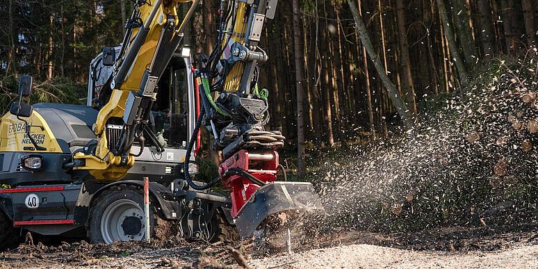 Kolové rypadlo Wacker Neuson EW100, které na lesní mýtině obsluhuje drtič pařezů, drtí pařez a rozmetává dřevní štěpku do vzduchu, v pozadí jsou naskládané klády a lesní porost.