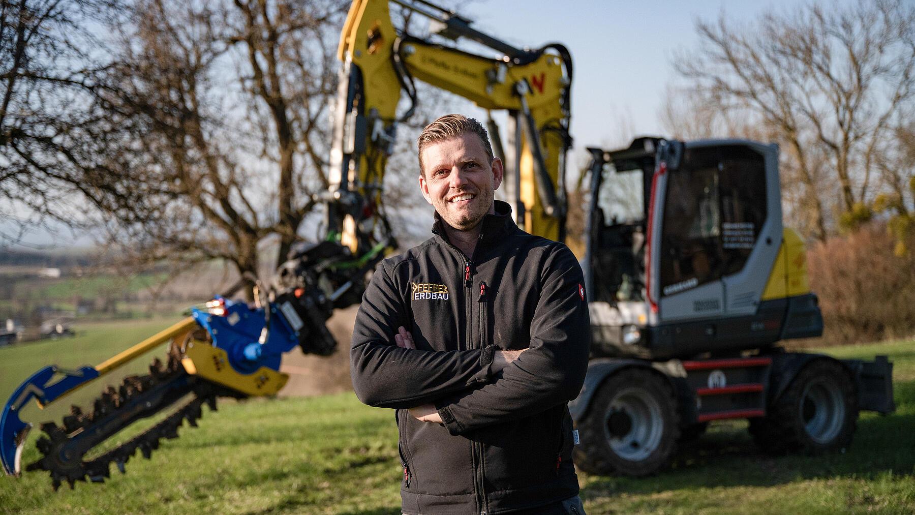 Contractor standing in front of a Wacker Neuson EW100 wheeled excavator fitted with a trenching attachment in a rural work area.