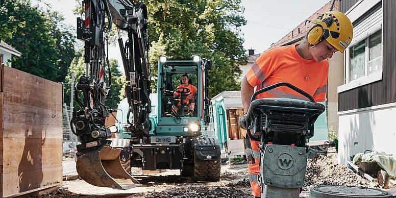 Stavební dělník obsluhuje pěch Wacker Neuson, zatímco v pozadí pracuje rypadlo EW100.