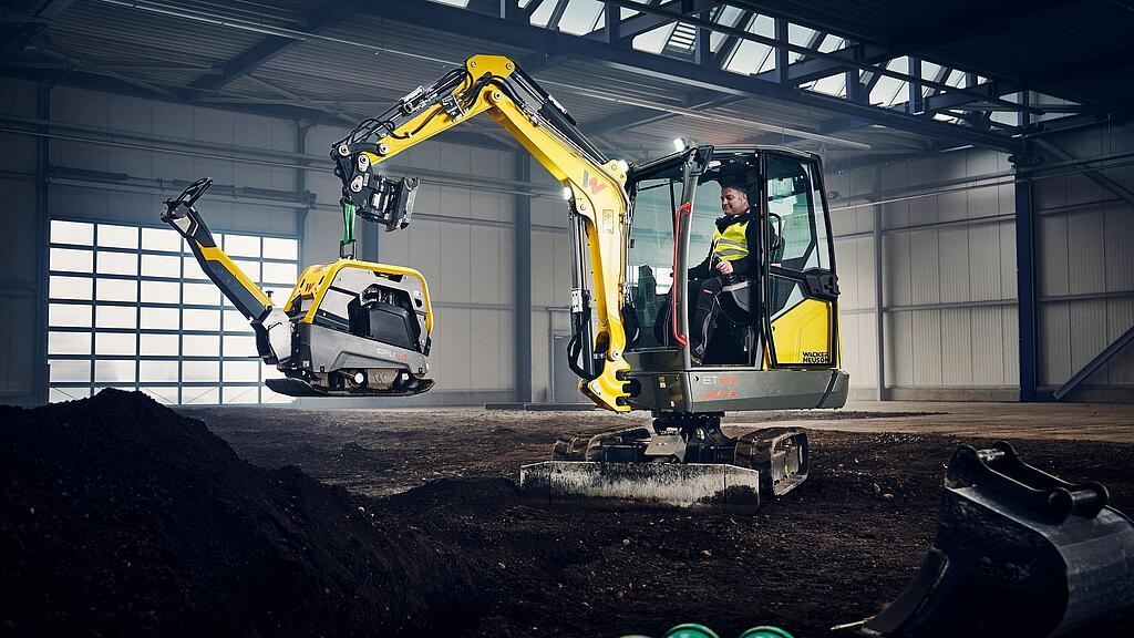 Wacker Neuson Minibagger ET25 hebt Rüttelplatte DPU62 in Industriehalle