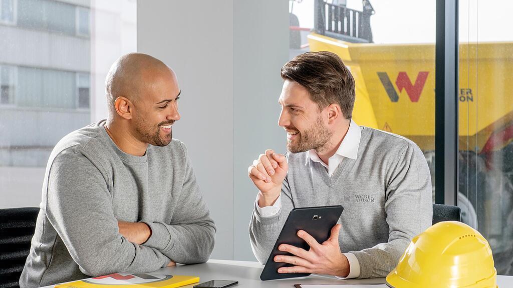 Zaměstnanec Wacker Neuson sedí u psacího stolu a ukazuje zákazníkům něco na svém tabletu.