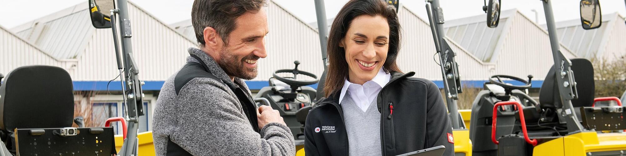 Usměvavý zaměstnanec Wacker Neuson s tabletem radí usmívajícímu se zákazníkovi před stavebními stroji Wacker Neuson.